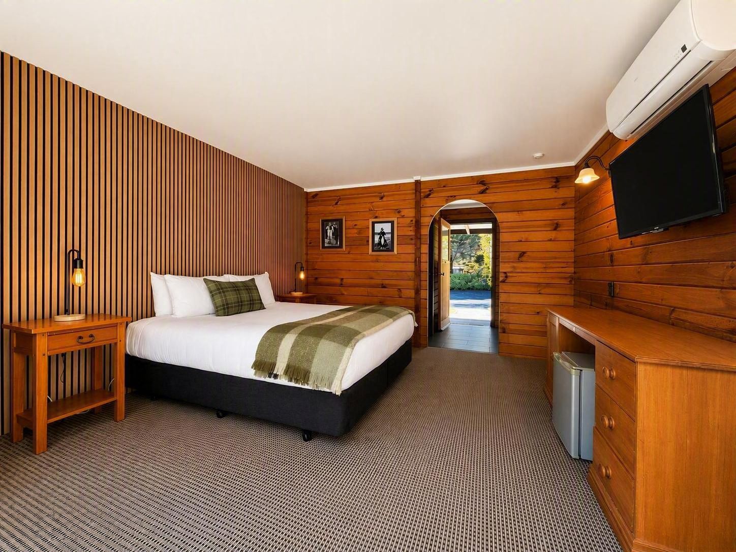 A hotel room with a bed, bedside table, lamp, TV, and a view through an arched doorway.
