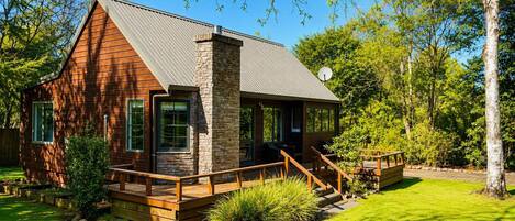 A cabin with a stone chimney, wooden deck, and a covered outdoor seating area.