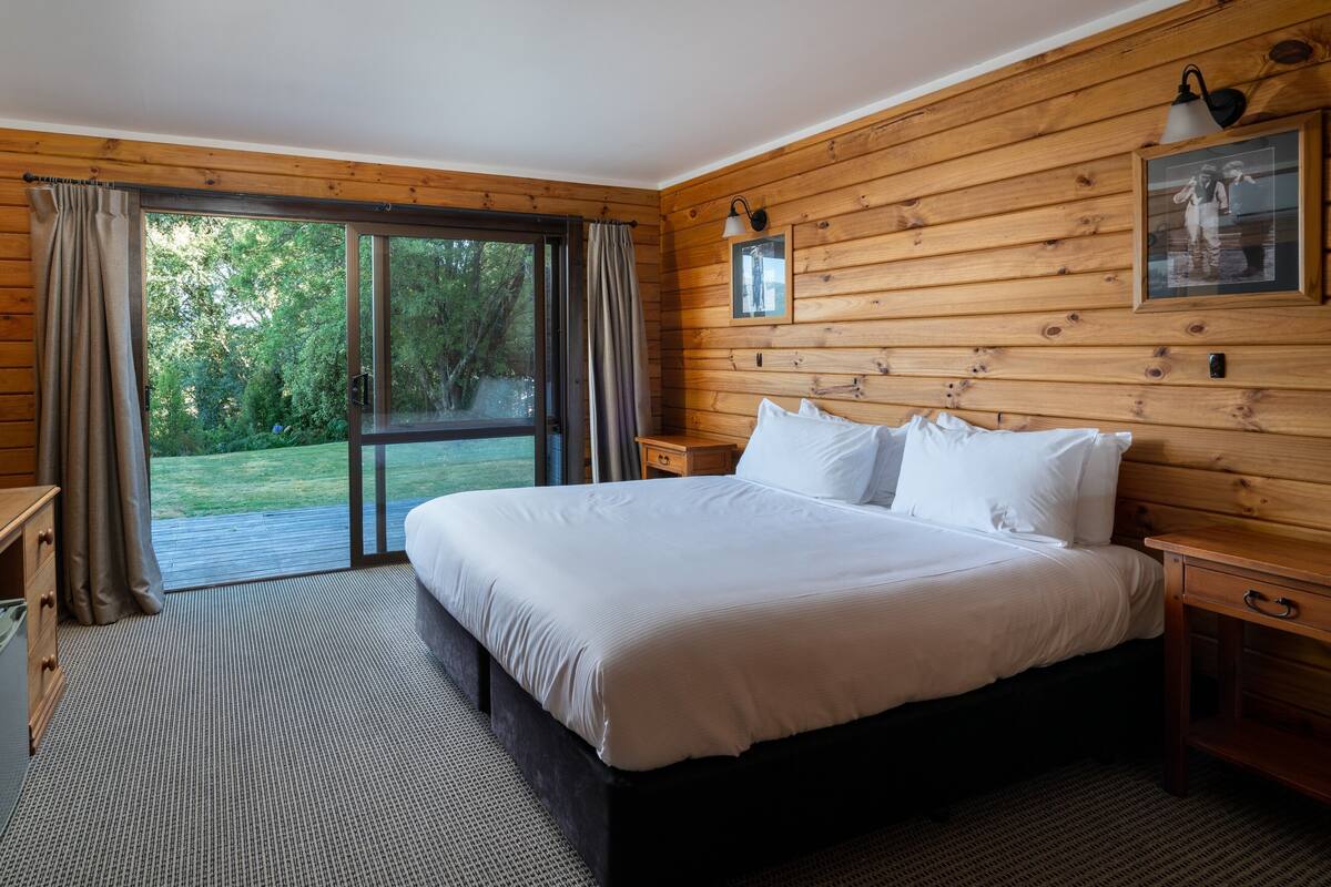 A bedroom with a large bed, wooden walls, a sliding glass door to a balcony, and a framed picture on the wall.