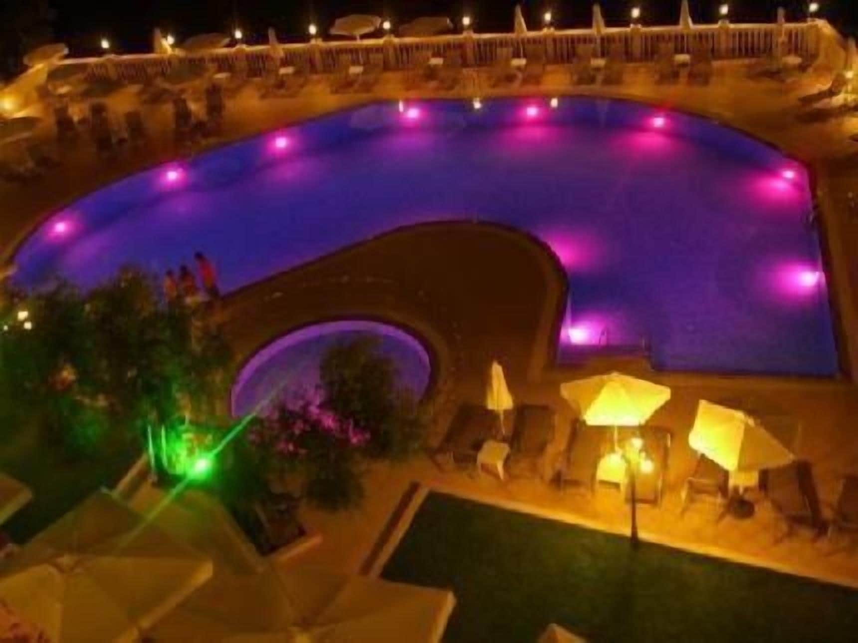 A nighttime view of a curved swimming pool with surrounding lounge chairs and umbrellas, illuminated by purple and green lights.