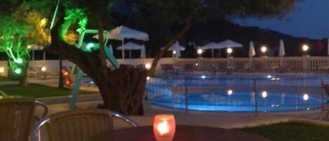 Zona de piscina al aire libre con mesa y sillas, rodeada de árboles y sombrillas.