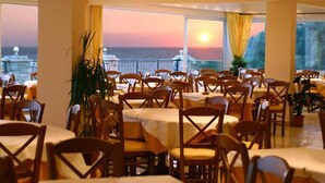 Un restaurante con mesas y sillas dispuestas para comer, amplias ventanas que ofrecen vista al mar y un balcón con barandilla.