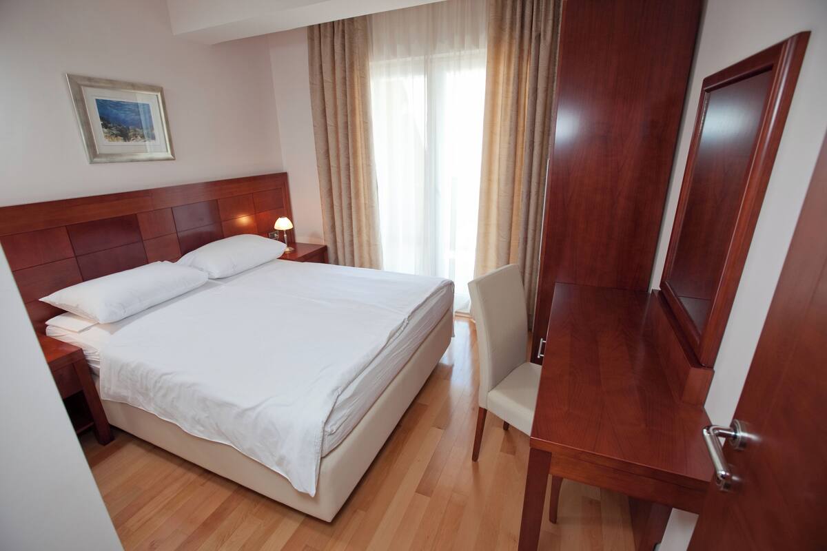 A hotel room with a bed, bedside table, lamp, chair, and a wooden desk.