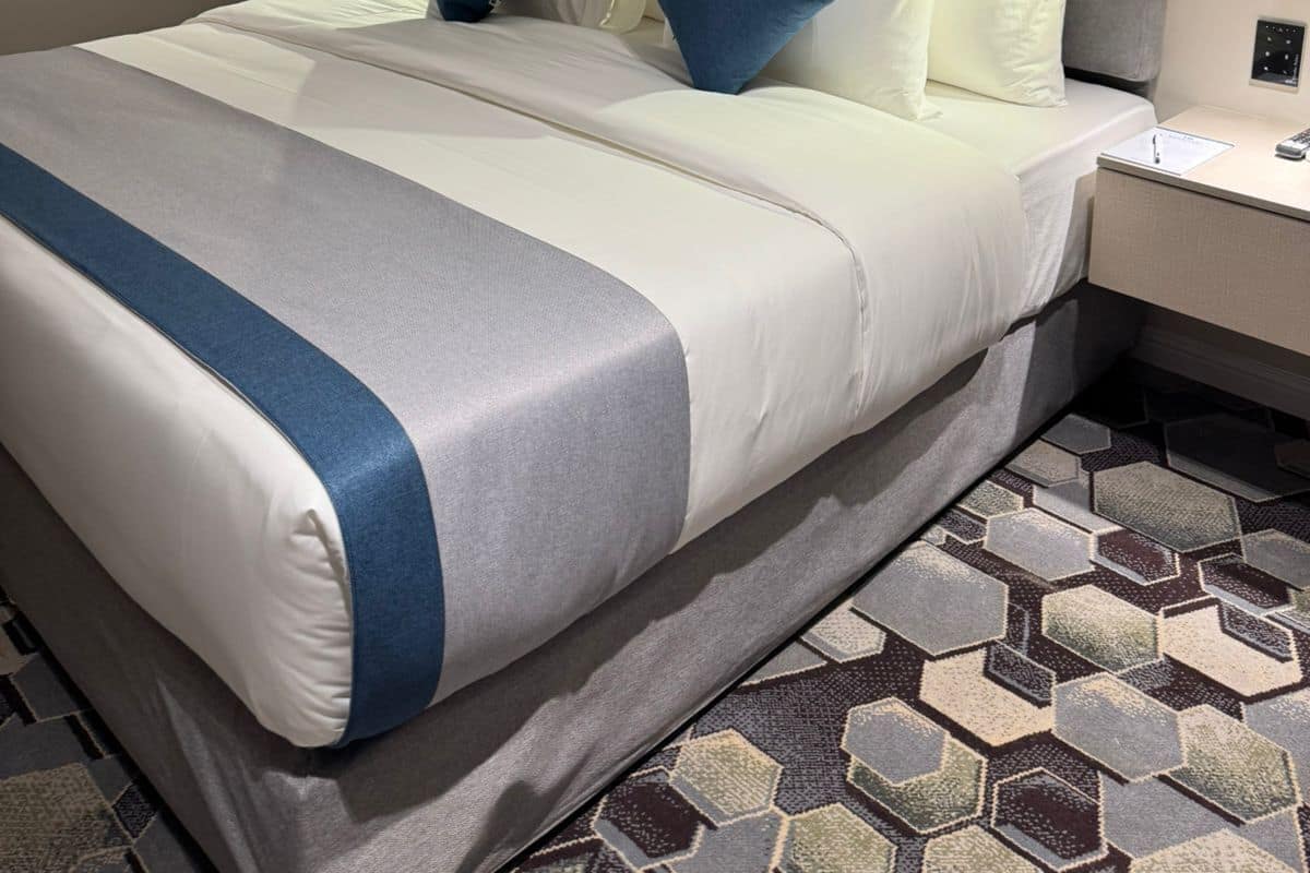 A modern hotel room with a large bed, a headboard, a bedside table, a nightstand, a wall-mounted artwork, and a patterned rug.