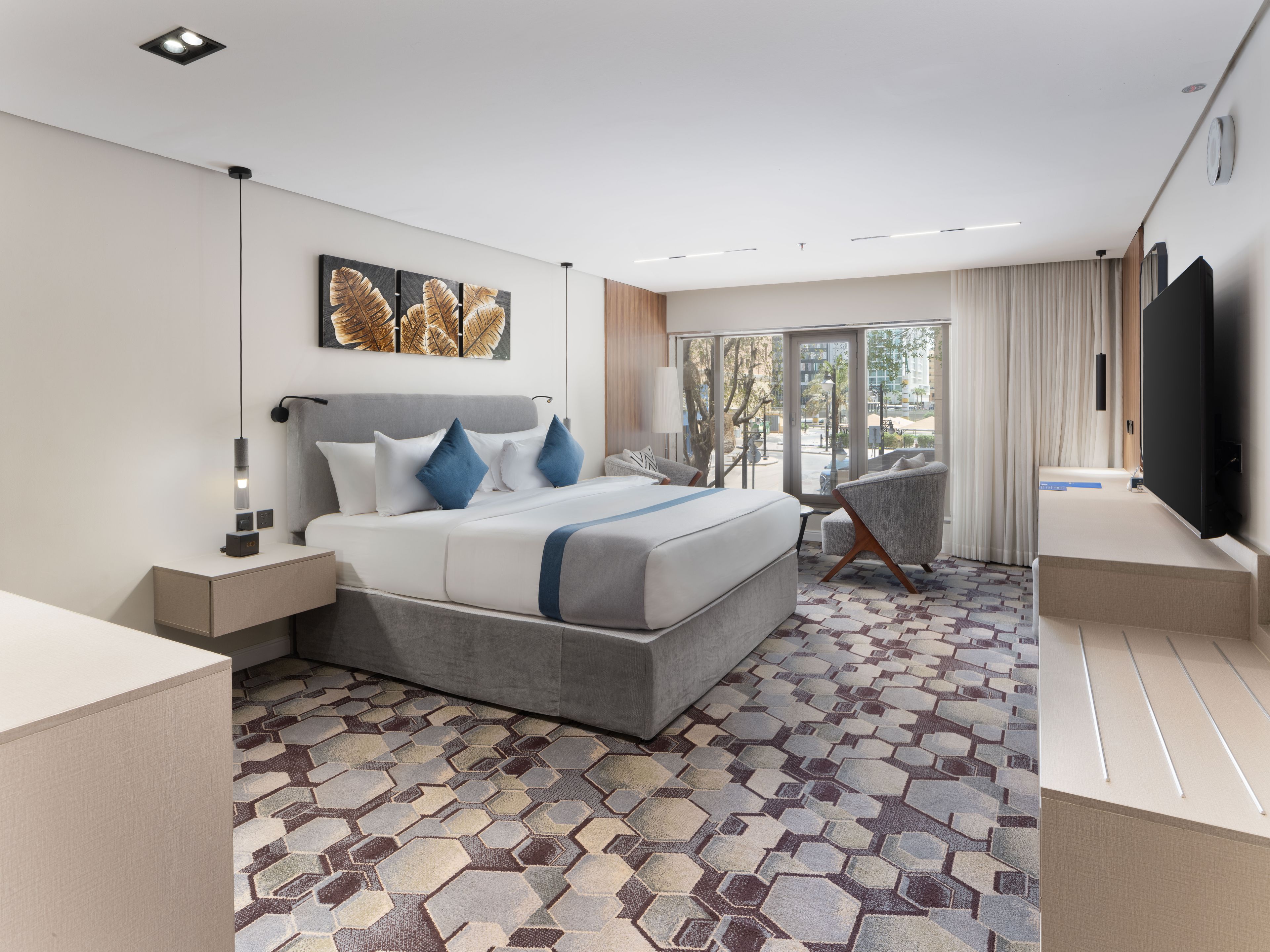 A modern hotel room with a large bed, a desk, a chair, a TV, and a view of the outdoors.