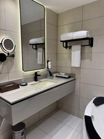 A modern bathroom with a large mirror, a sink, a towel rack, and a trash bin.