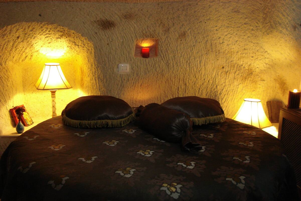 A bedroom with a stone wall, a bed with a floral bedspread, two pillows, a bedside table with a lamp, and a small vase.