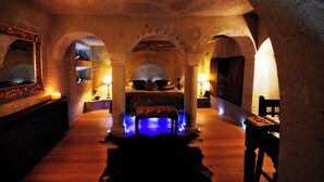 A dimly lit room with a bed, a bench, and a table with a lamp. - Perimasali Cave Hotel - Cappadocia (Ürgüp)