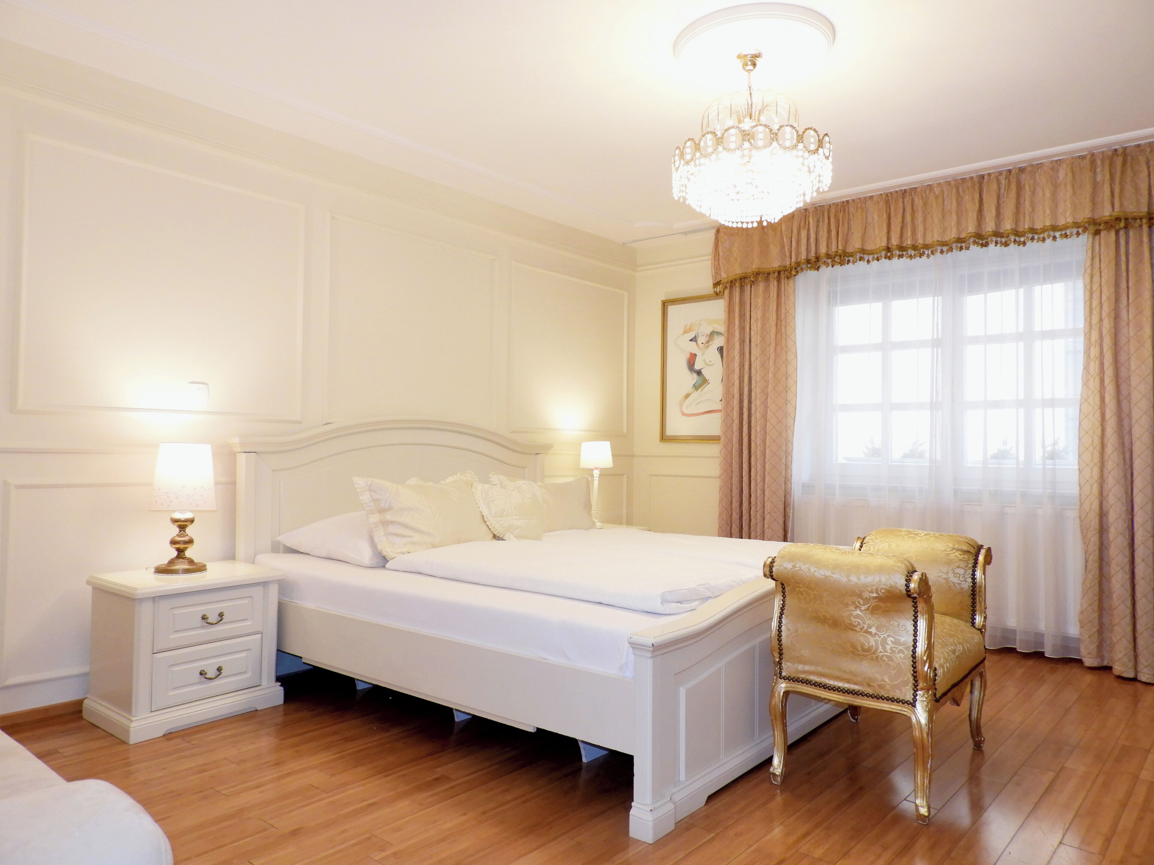 A bedroom with a bed, bedside tables, a chandelier, a chair, and a window with curtains.