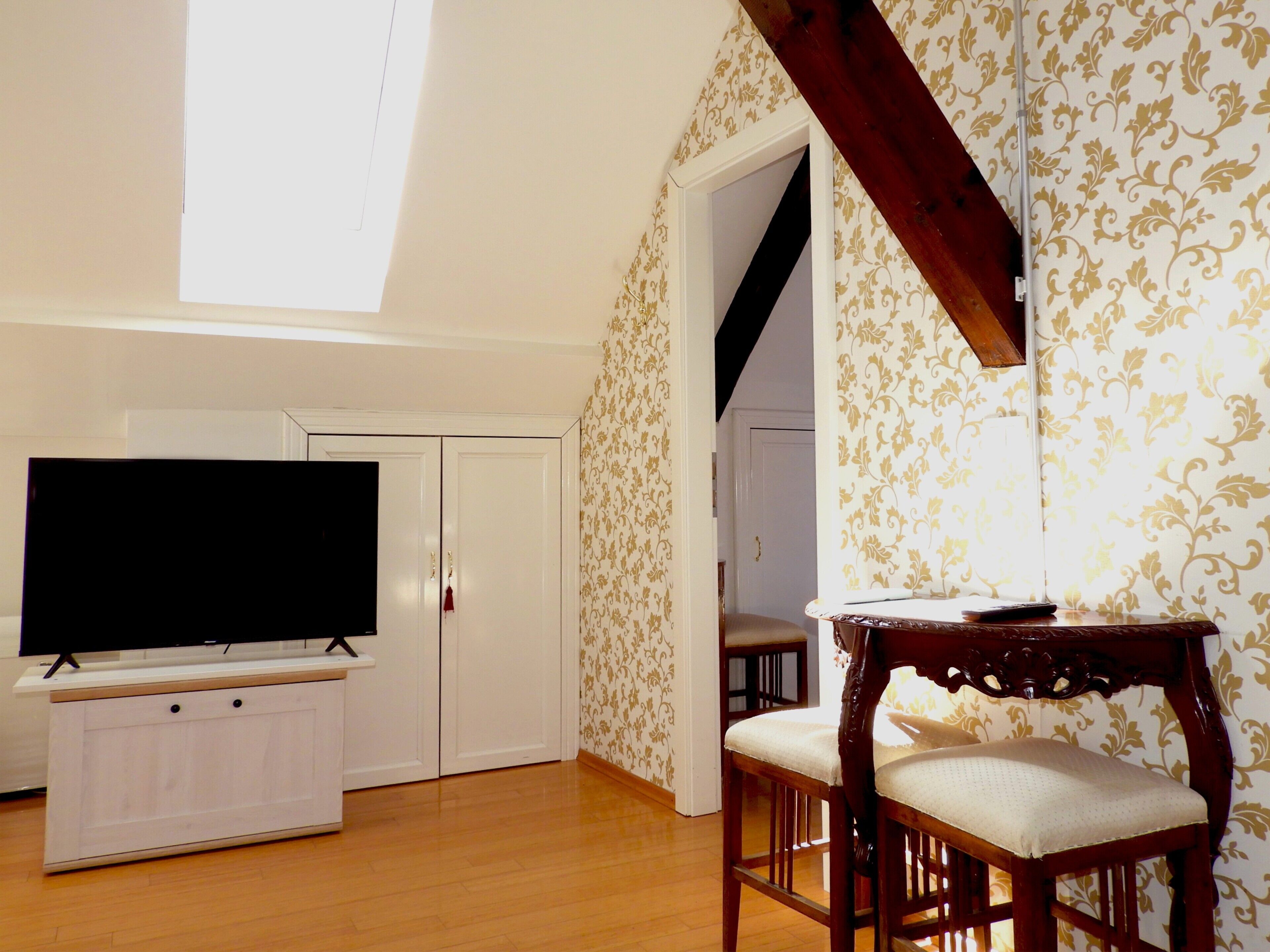 A room with a TV on a stand, a wooden chair, a wardrobe, and a patterned wallpaper.