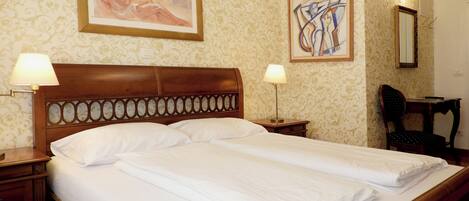 Una cama bien hecha con dos almohadas, un cabecero de madera, dos lámparas de noche, una silla y dos obras de arte enmarcadas en la pared.