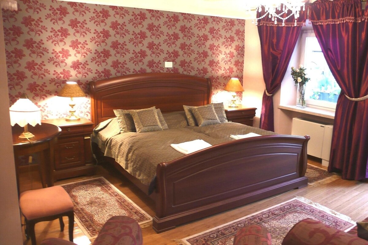A bedroom with a wooden bed, two bedside tables with lamps, a window with purple curtains, and a floral wallpaper.