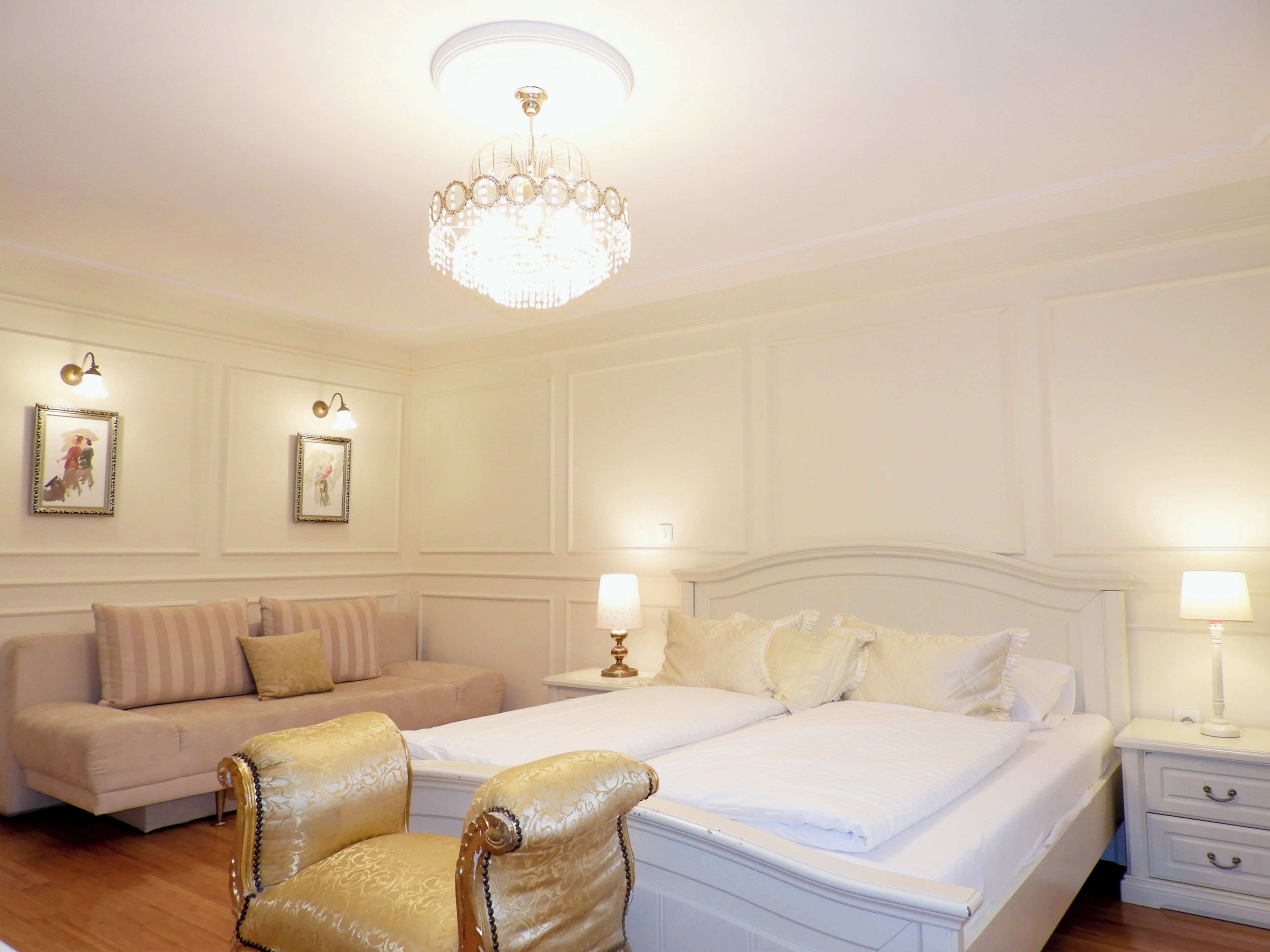 A hotel room with a large bed, two bedside tables with lamps, a sofa, a chair, and a chandelier.
