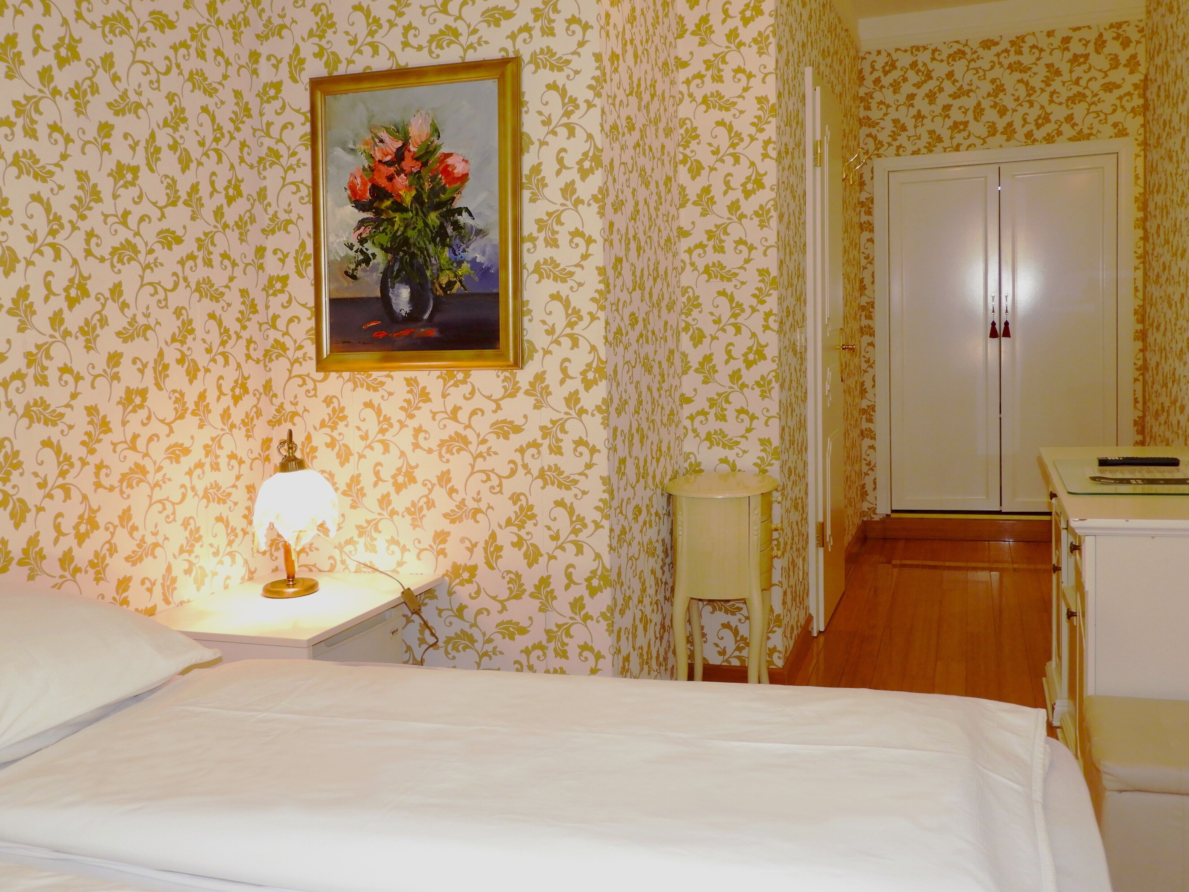 A hotel room with a bed, a bedside table with a lamp, a small stool, a door, and a painting on the wall.