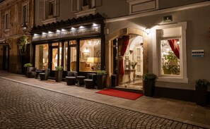 A hotel facade with a dark entrance, multiple windows, and outdoor seating with potted plants.