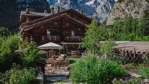 A wooden chalet with outdoor seating, surrounded by lush greenery and mountains in the background.