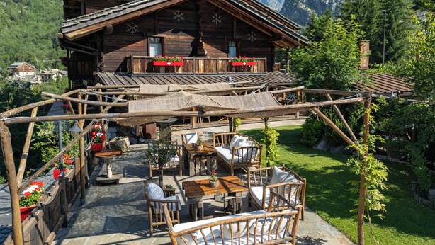 A wooden chalet with a balcony, outdoor seating, and a view of snow-capped mountains.