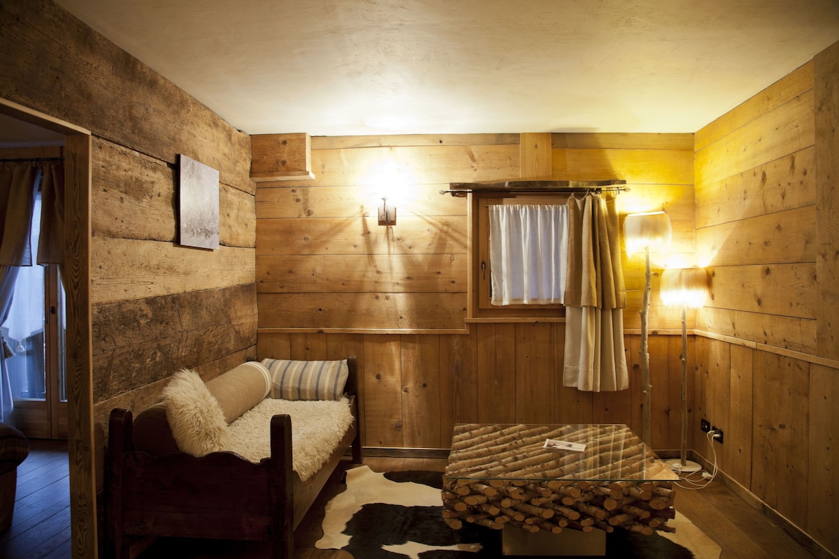 A cozy cabin-style room with wooden walls, a bed with a fur throw, a small table with logs, and a window with curtains.