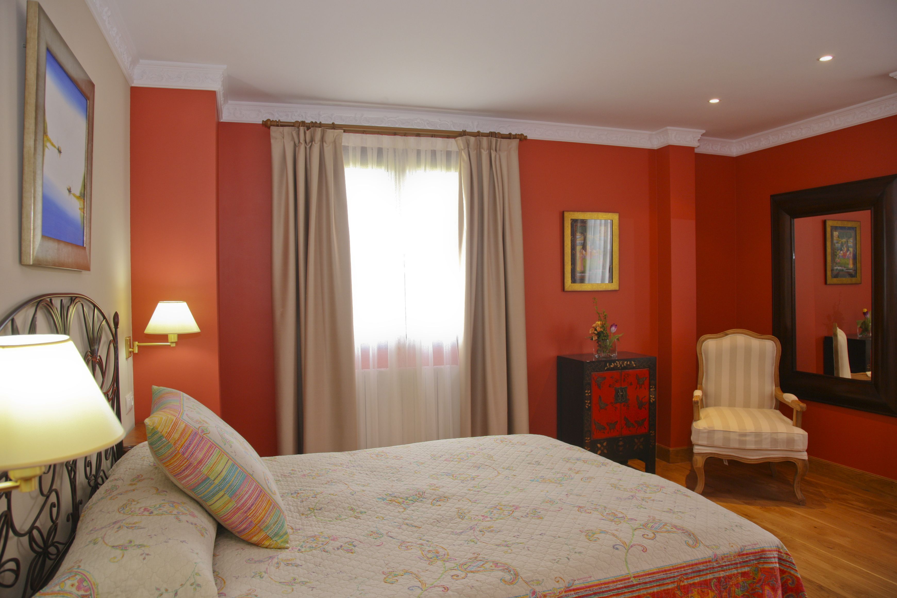 Una habitación de hotel con cama, una silla, una lámpara, ventana con cortinas y un cuadro en la pared.
