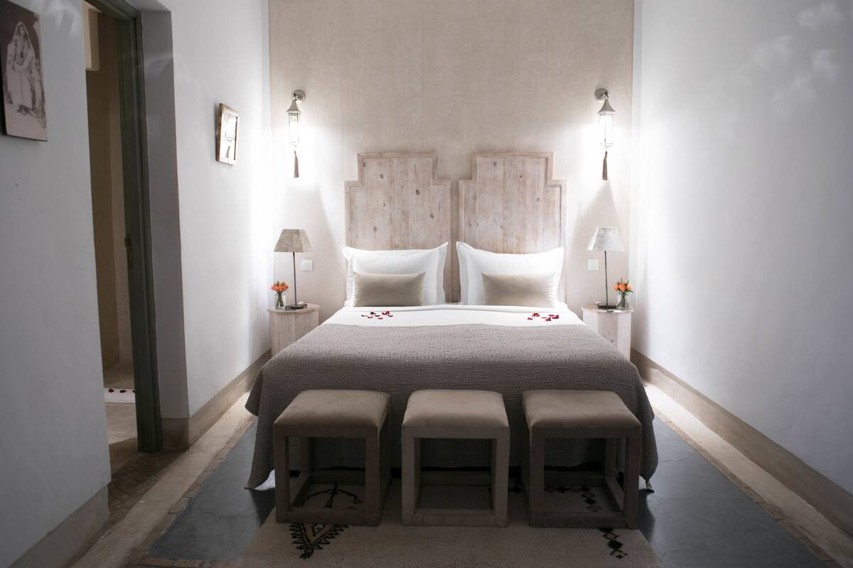 Un dormitorio con una cama grande, dos mesitas de noche, una lámpara de pared y un taburete pequeño.