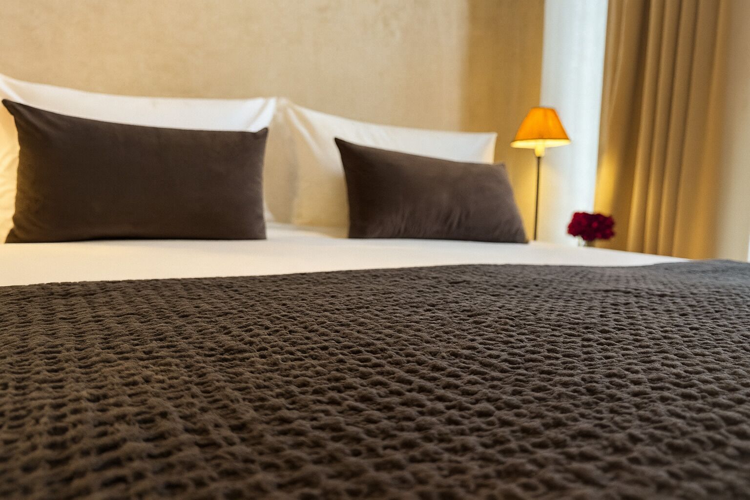 A bed with a textured blanket, two pillows, a lamp, and a vase with a flower.