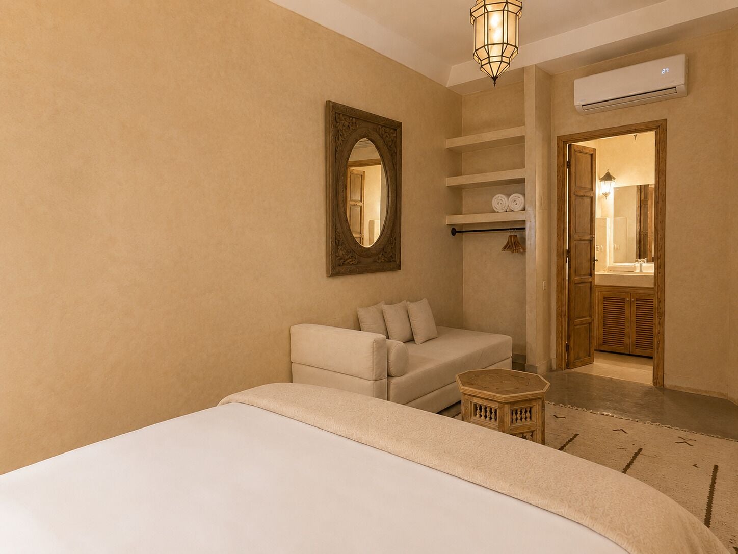A hotel room with a bed, a seating area with a chair and a small table, a mirror, a shelf with towels, and an air conditioning unit.