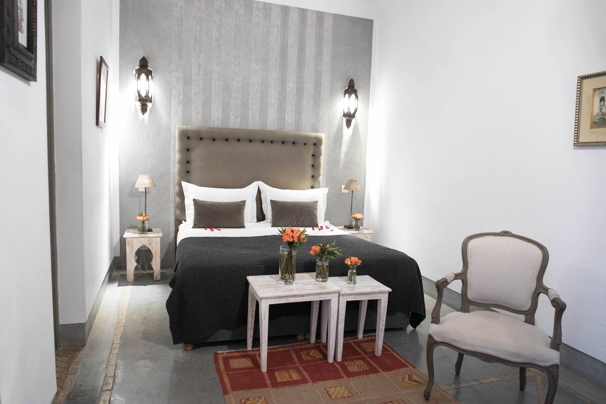 Un dormitorio con una cama grande, dos mesitas de noche con lámparas, una silla y una pequeña mesa con jarrones de flores.