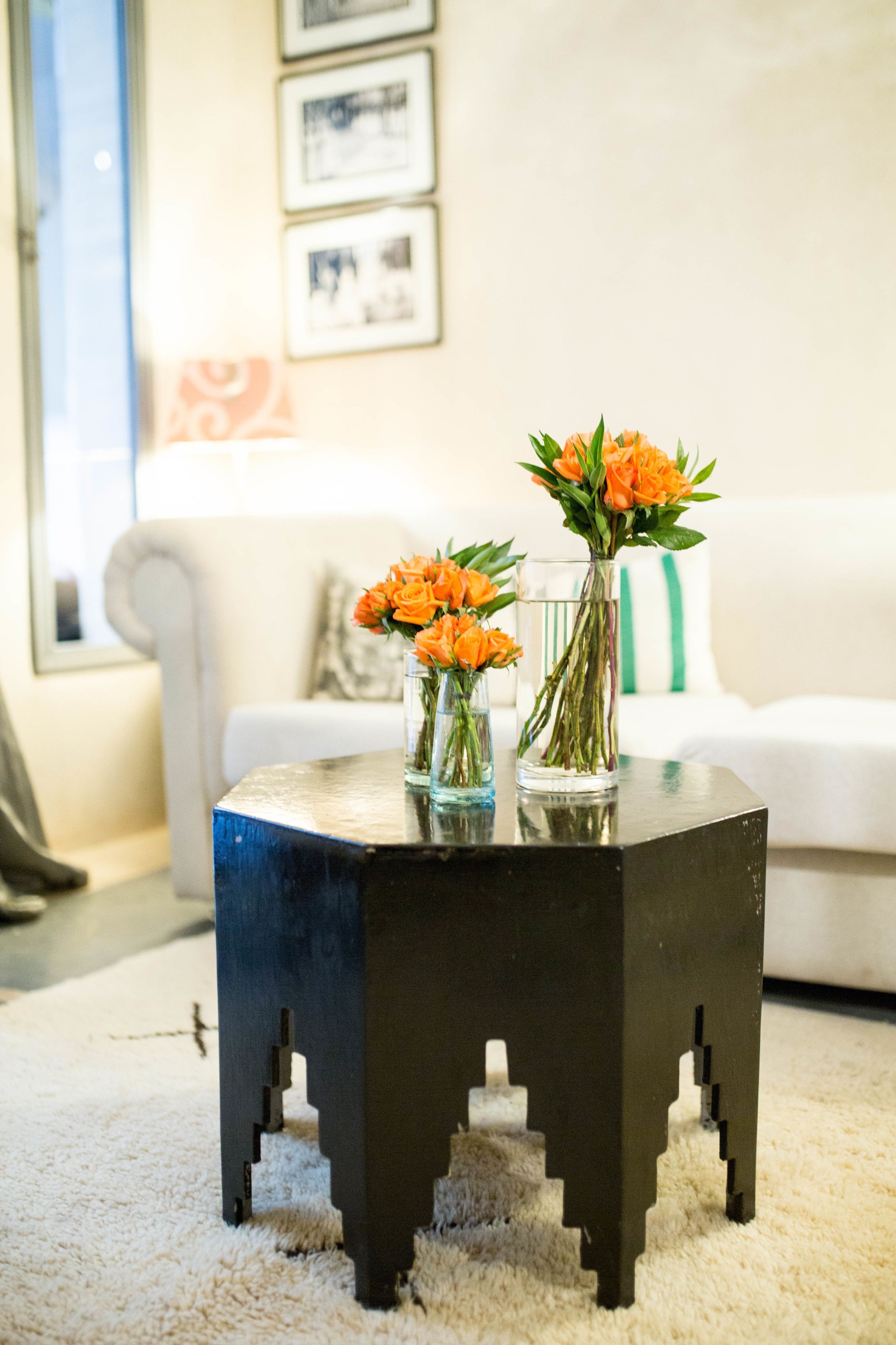 A black side table with two vases of orange flowers on a white carpeted floor.