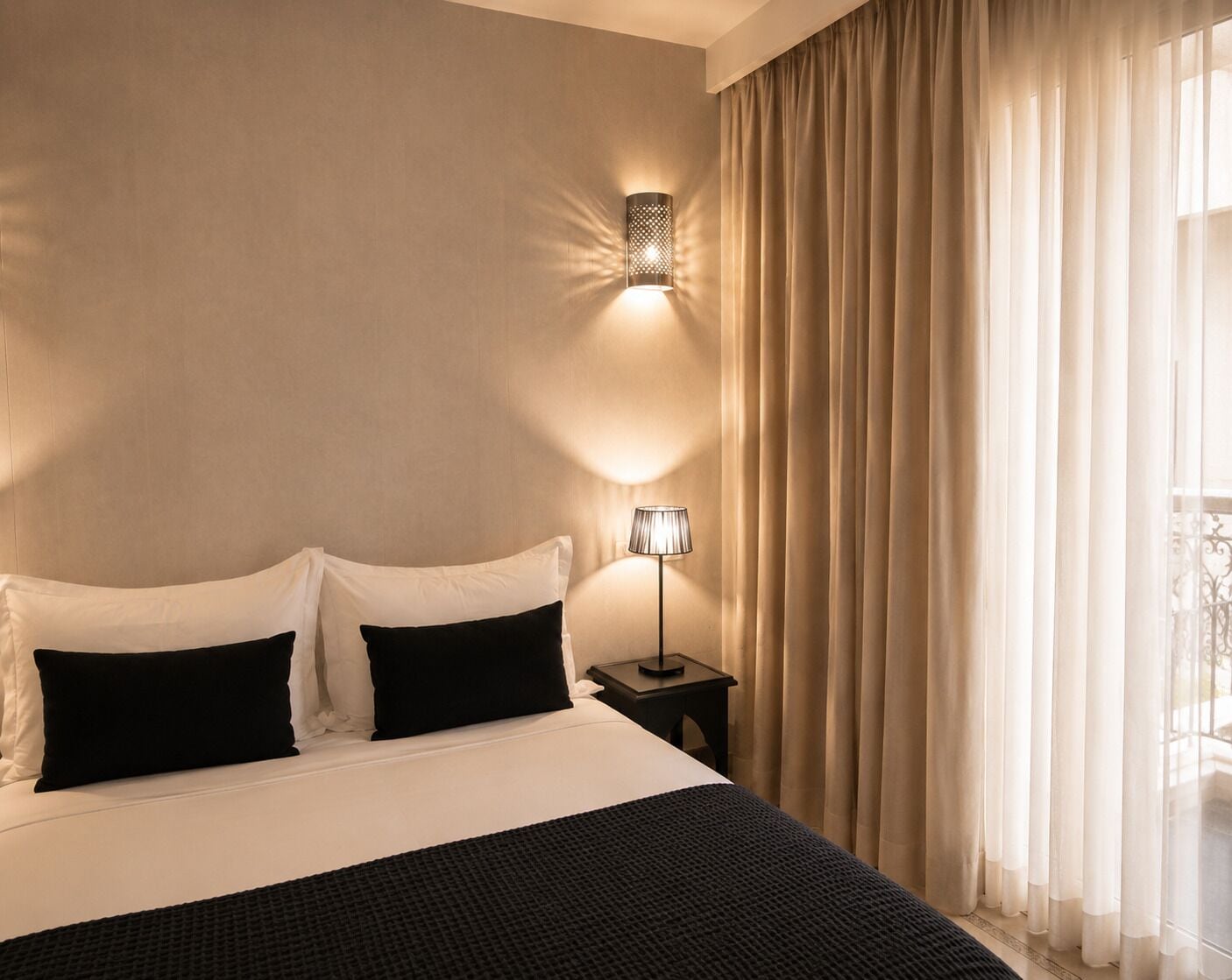 Een hotelkamer met een bed, twee kussens, een nachtkastje met een lamp, en een raam met gordijnen.
