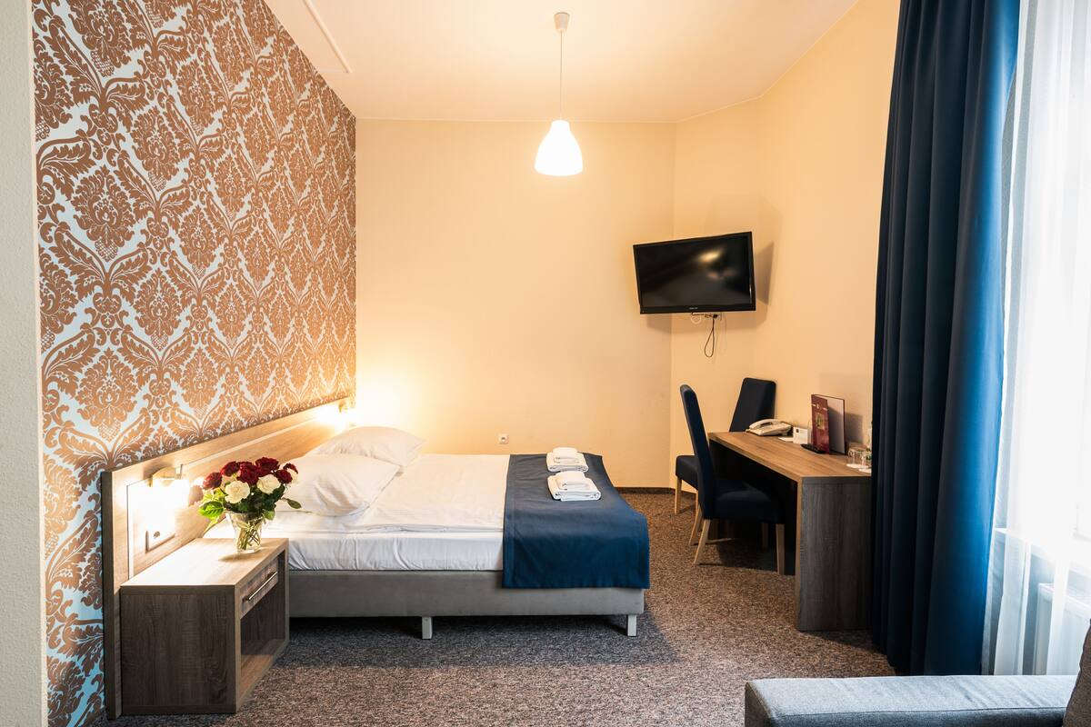 A hotel room with a bed, bedside table with a lamp, a desk with a chair, a TV mounted on the wall, and a window with curtains.