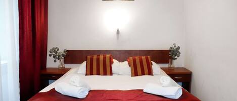 Habitación de hotel con una cama grande, ropa de cama roja, dos mesitas de noche con jarrones y un cuadro en la pared.