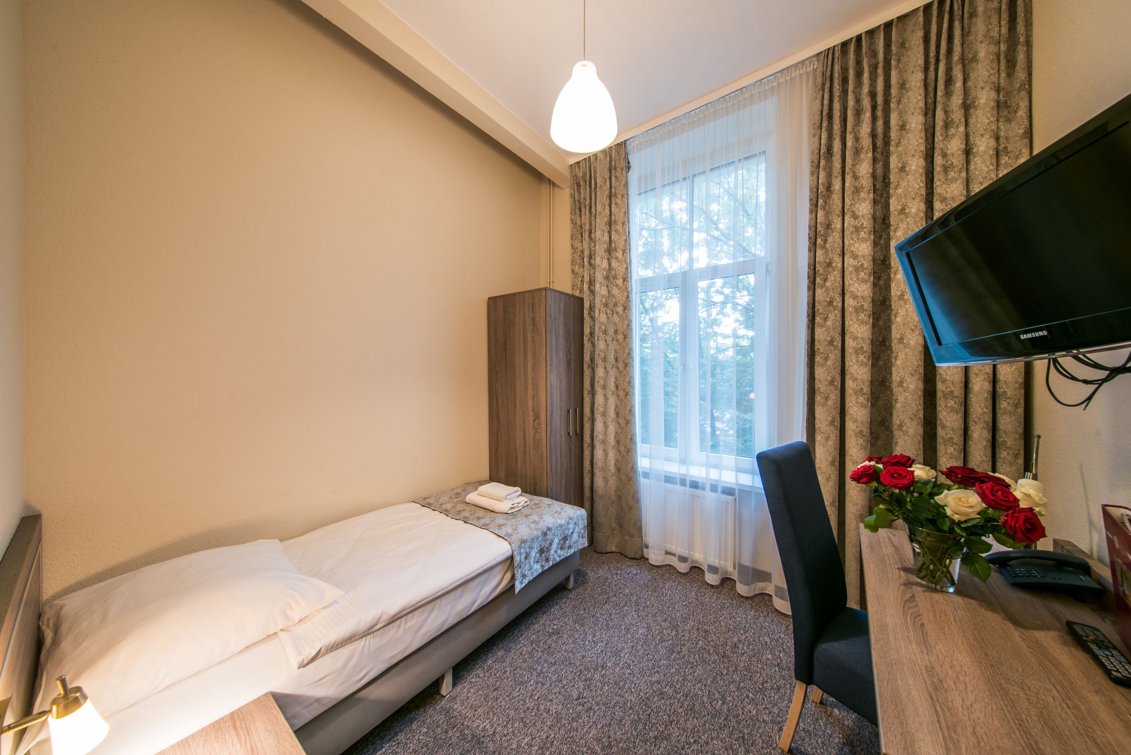 A small bedroom with a single bed, a desk with a computer, a television, and a vase of flowers.