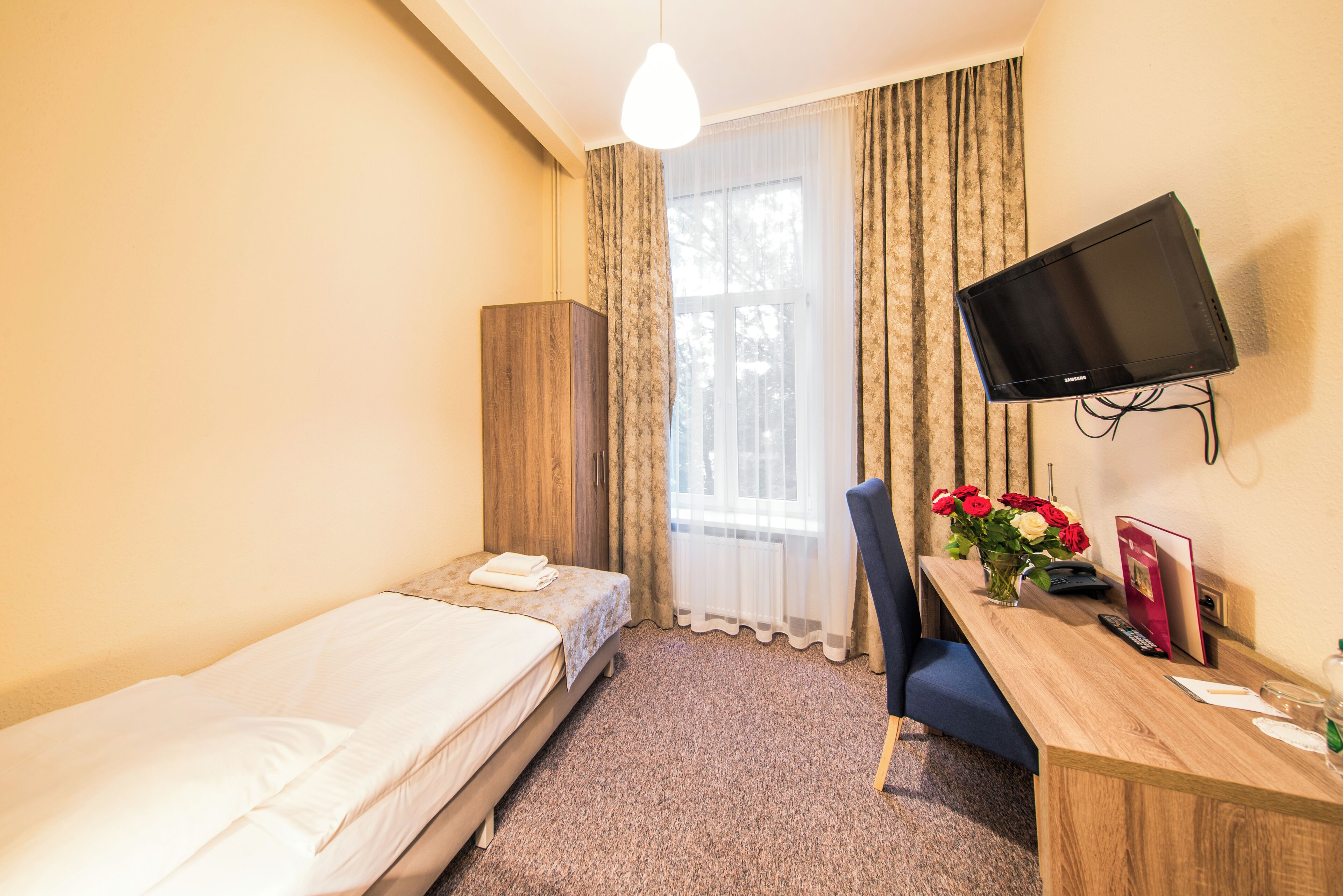 A hotel room with a bed, a desk with a chair, a television mounted on the wall, and a window with curtains.