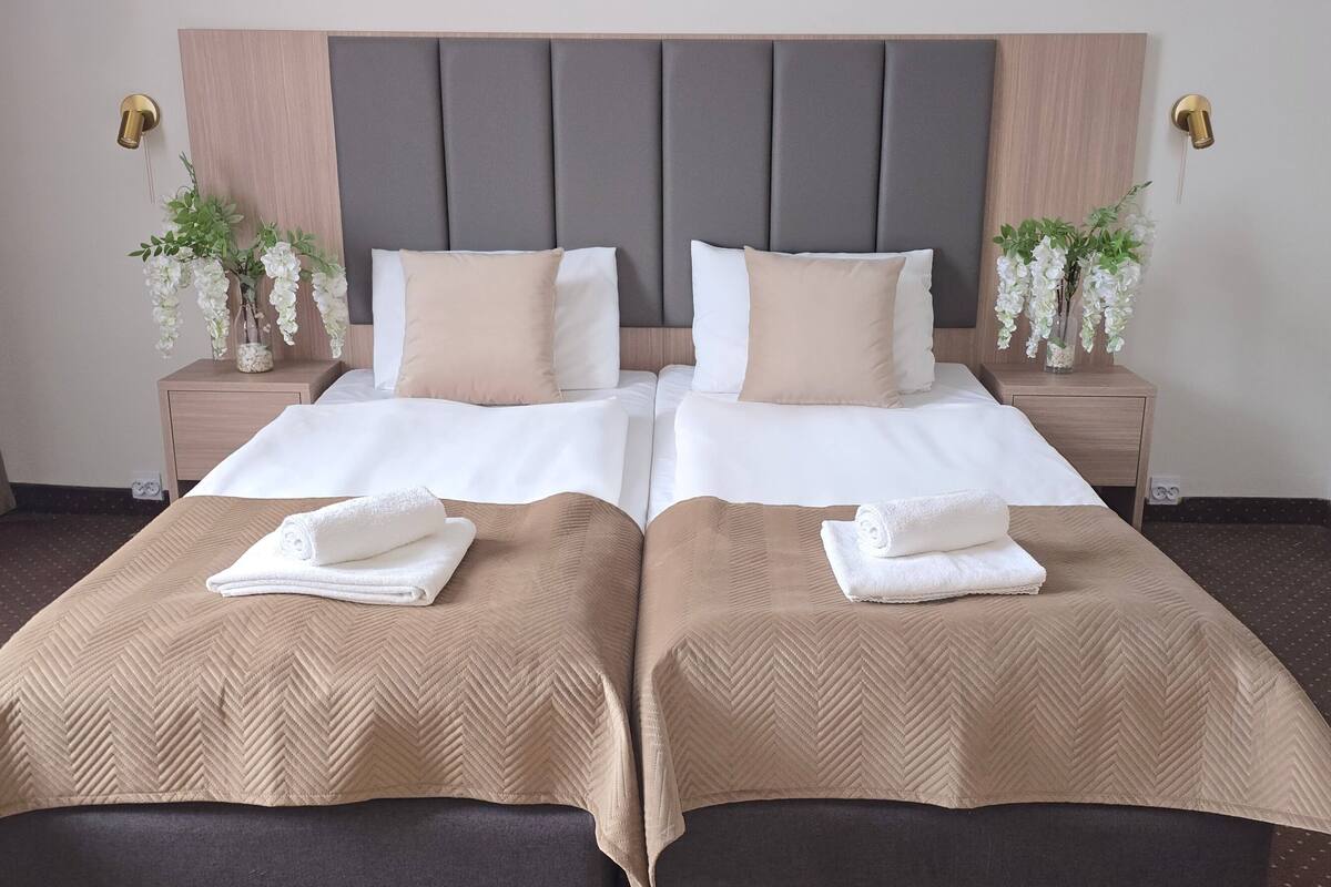 A double room with two beds, each with a beige bedspread and white pillows, flanked by nightstands with white flowers.