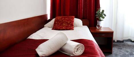 A neatly made bed with a red and white bedspread, a rolled white towel, and a red pillow with a gold pattern.