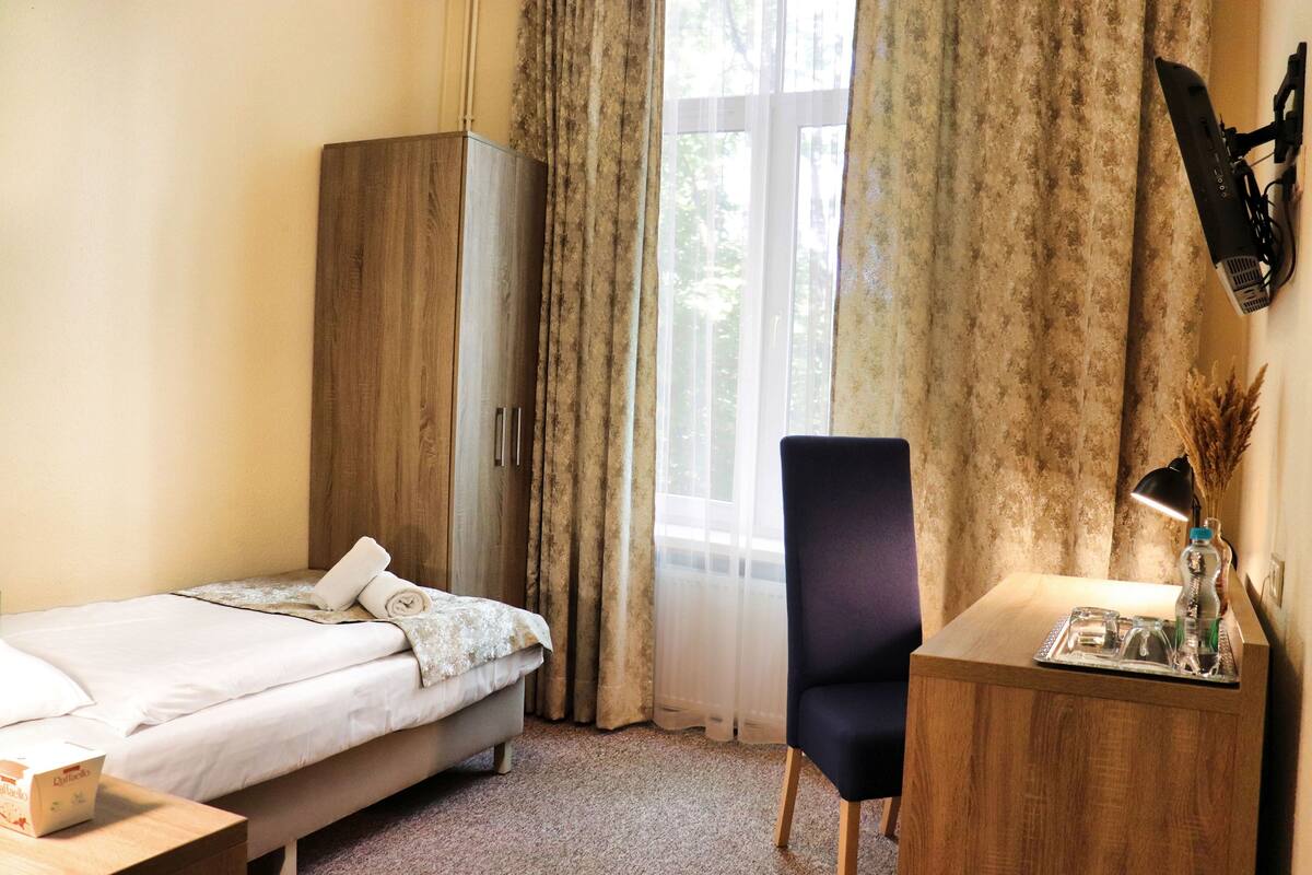 A hotel room with a bed, a desk with a lamp, a chair, a wardrobe, and a window with curtains.