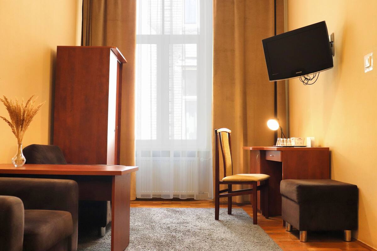 A room with a wooden desk, a chair, a sofa, a TV mounted on the wall, and a window with curtains.