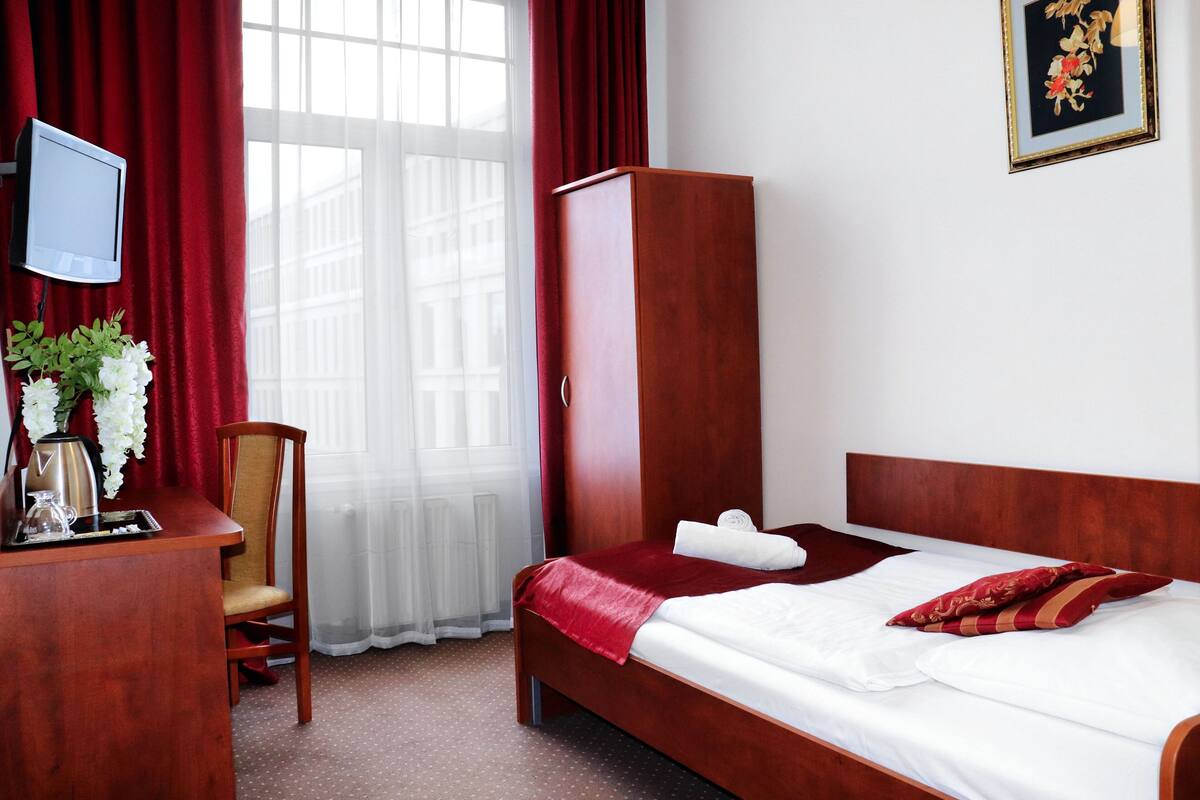 A hotel room with a bed, a wooden wardrobe, a desk with a chair, a TV mounted on the wall, and a window with red curtains.