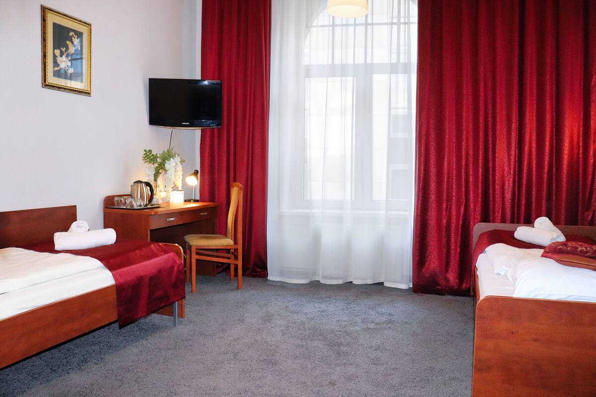 A hotel room with two beds, a desk with a chair, a television, and a large window with red curtains.