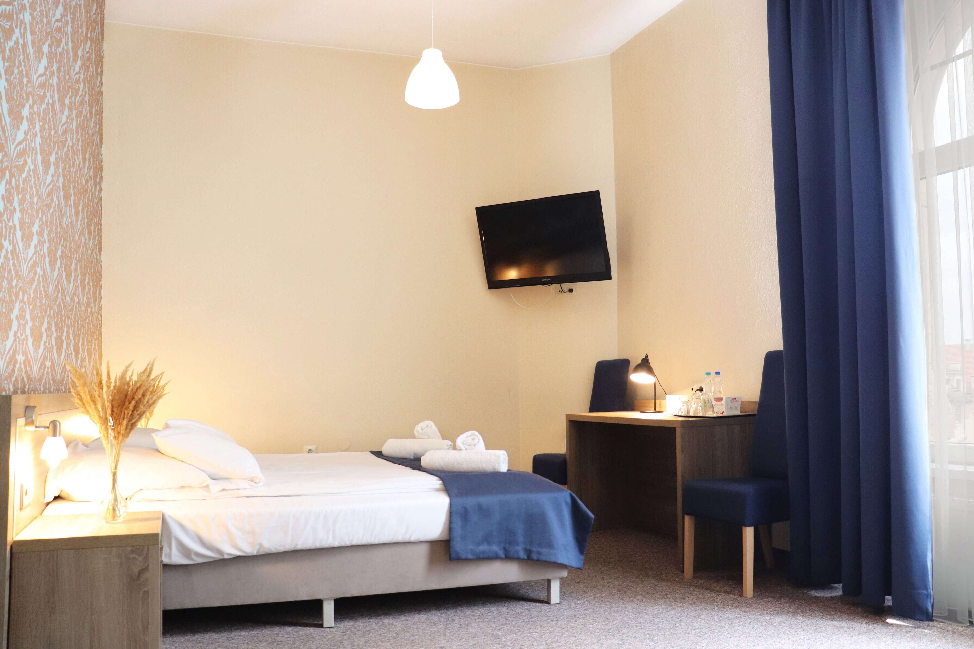 A hotel room with a bed, a desk, a chair, a TV mounted on the wall, and a window with curtains.