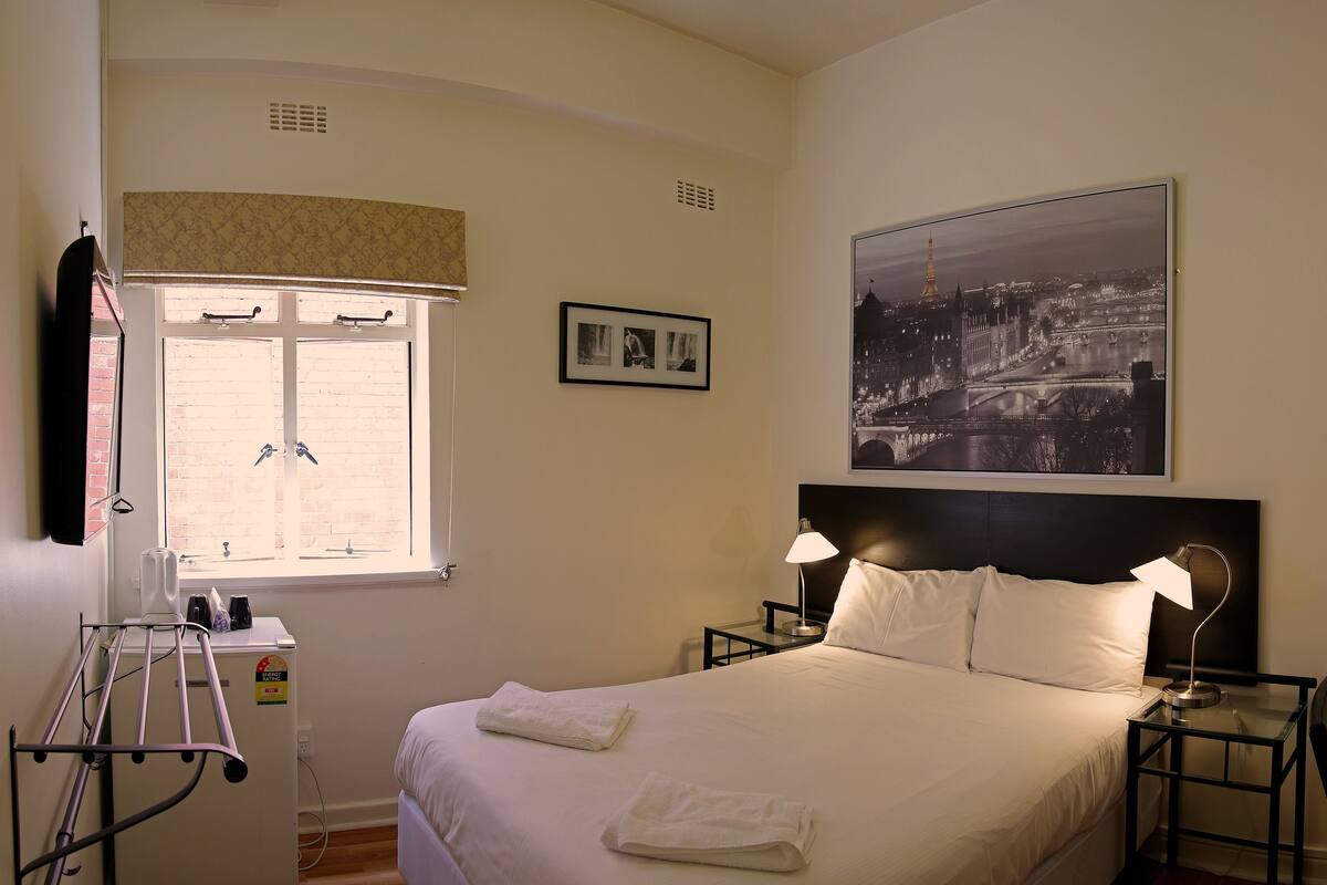 Une chambre d’hôtel bien rangée, avec un lit, une table de chevet, une table, une lampe, une fenêtre avec des stores, un cadre photo et un miroir fixé au mur.