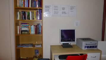 Una oficina pequeña con escritorio, computadora, impresora y una estantería llena de libros. Hay letreros en la pared con instrucciones para los huéspedes.