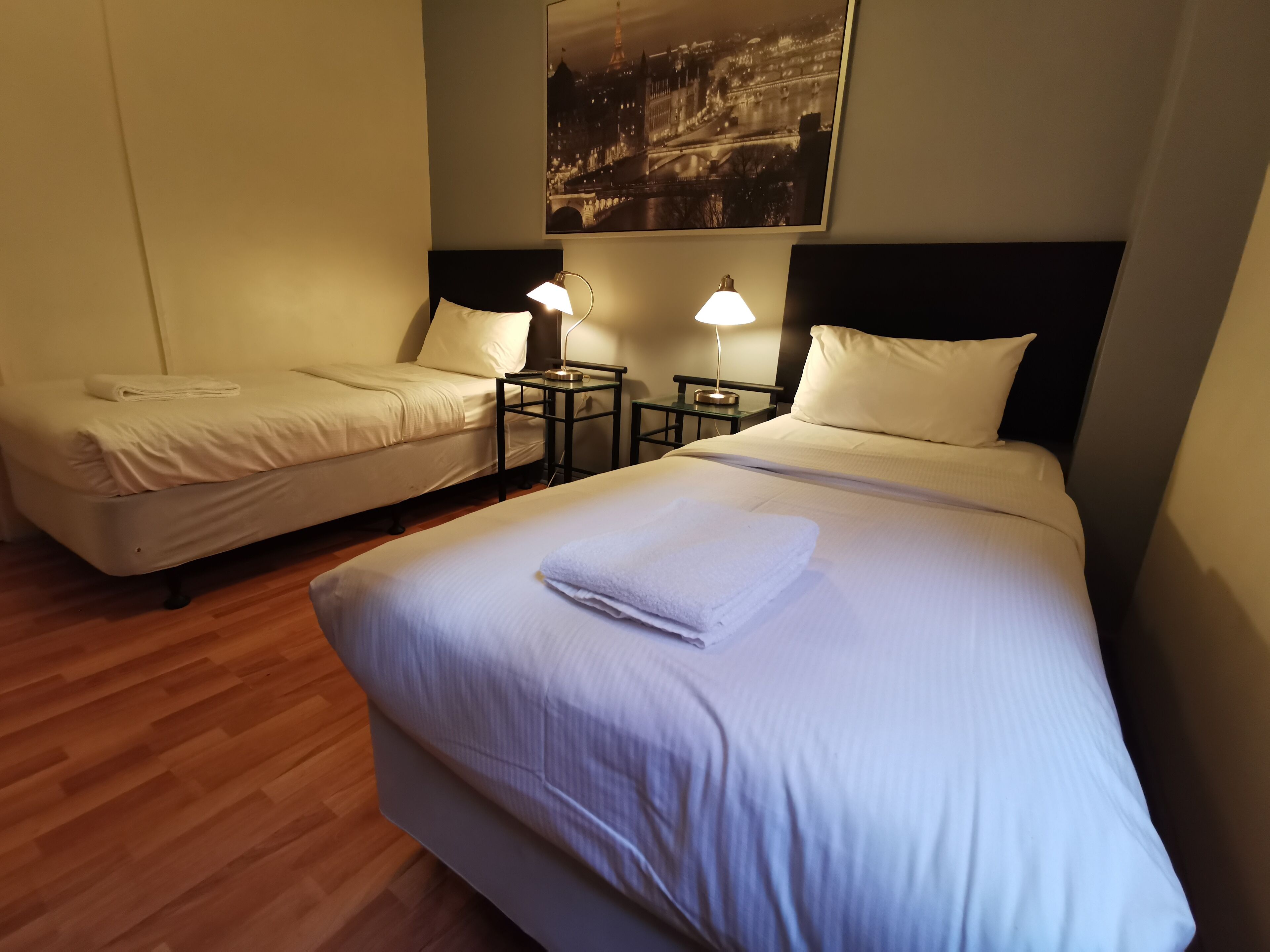 A hotel room with two single beds, a nightstand with a lamp, and a large bed with a folded towel.