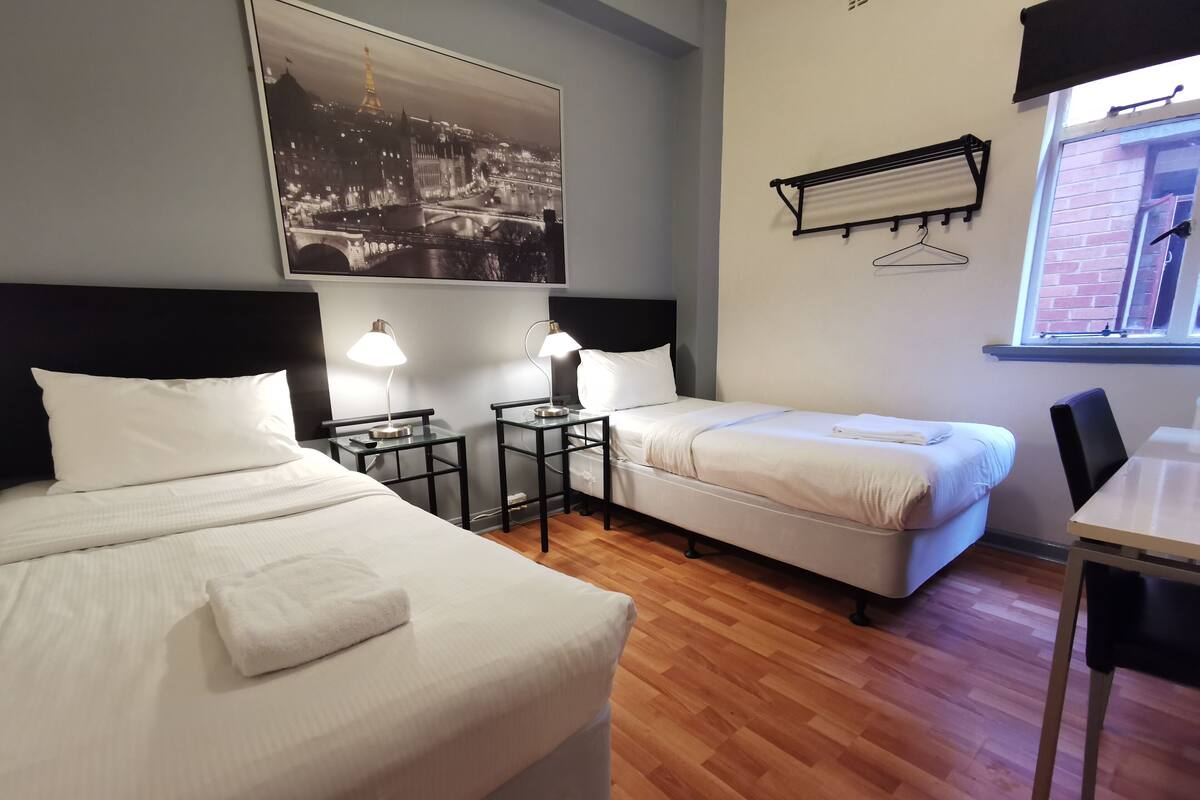 Une chambre d’hôtel avec deux lits, un bureau, une chaise, une fenêtre donnant sur un paysage urbain et un climatiseur fixé au mur.