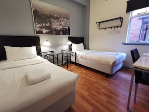 A hotel room with two beds, a desk, a chair, a window with a view of a cityscape, and a wall-mounted air conditioning unit. - City Centre Budget Hotel (Melbourne)