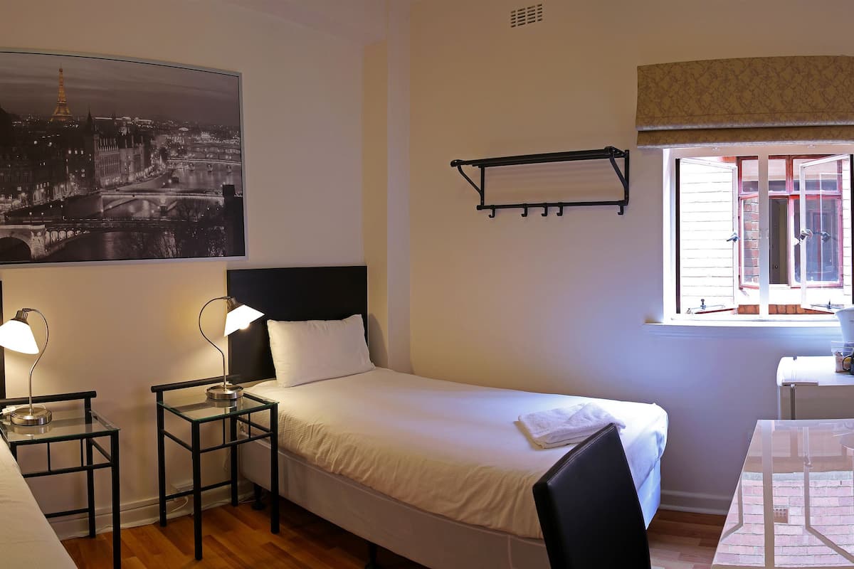 Une chambre d’hôtel avec deux lits simples, une petite table, une chaise, une fenêtre avec des volets, un porte-manteau et un téléviseur fixé au mur.