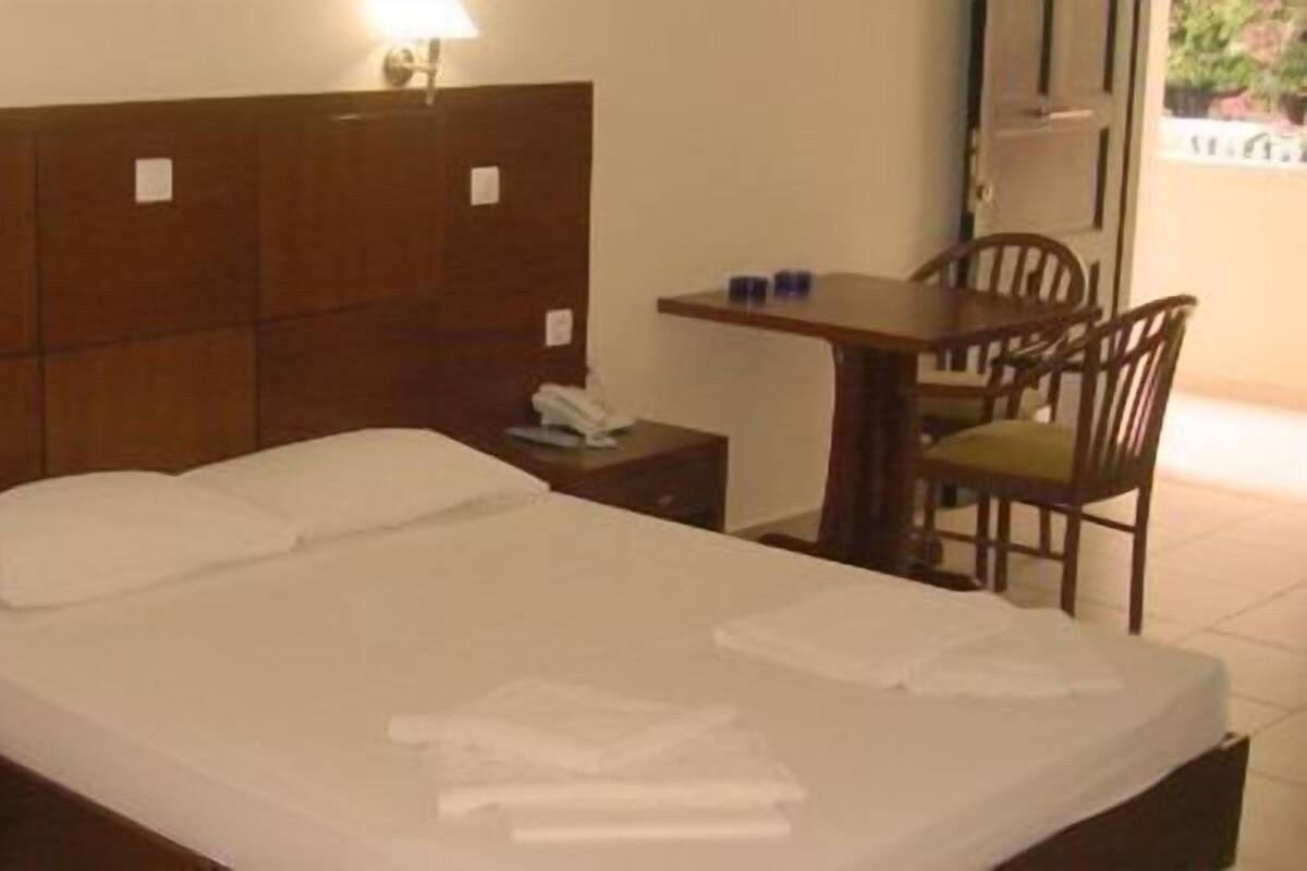 A hotel room with a wooden headboard, a single bed with white linens, a bedside table with a phone, a small desk with a chair, and a view of greenery outside.