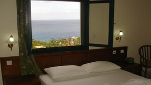 A hotel room with a bed, a nightstand, a lamp, a telephone, and a view of the ocean. - Kyra Panaghia (Karpathos)