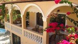 A covered outdoor seating area with tables and chairs, a pool, and a building with arches and a balcony. - Kyra Panaghia (Karpathos)
