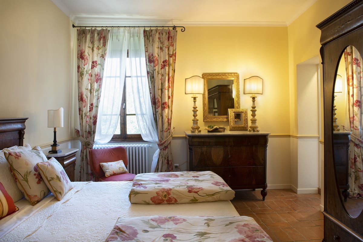 Ein Schlafzimmer mit einem großen Bett, zwei Nachttischen, einer Kommode, einem Spiegel und einem Fenster mit floralen Vorhängen.